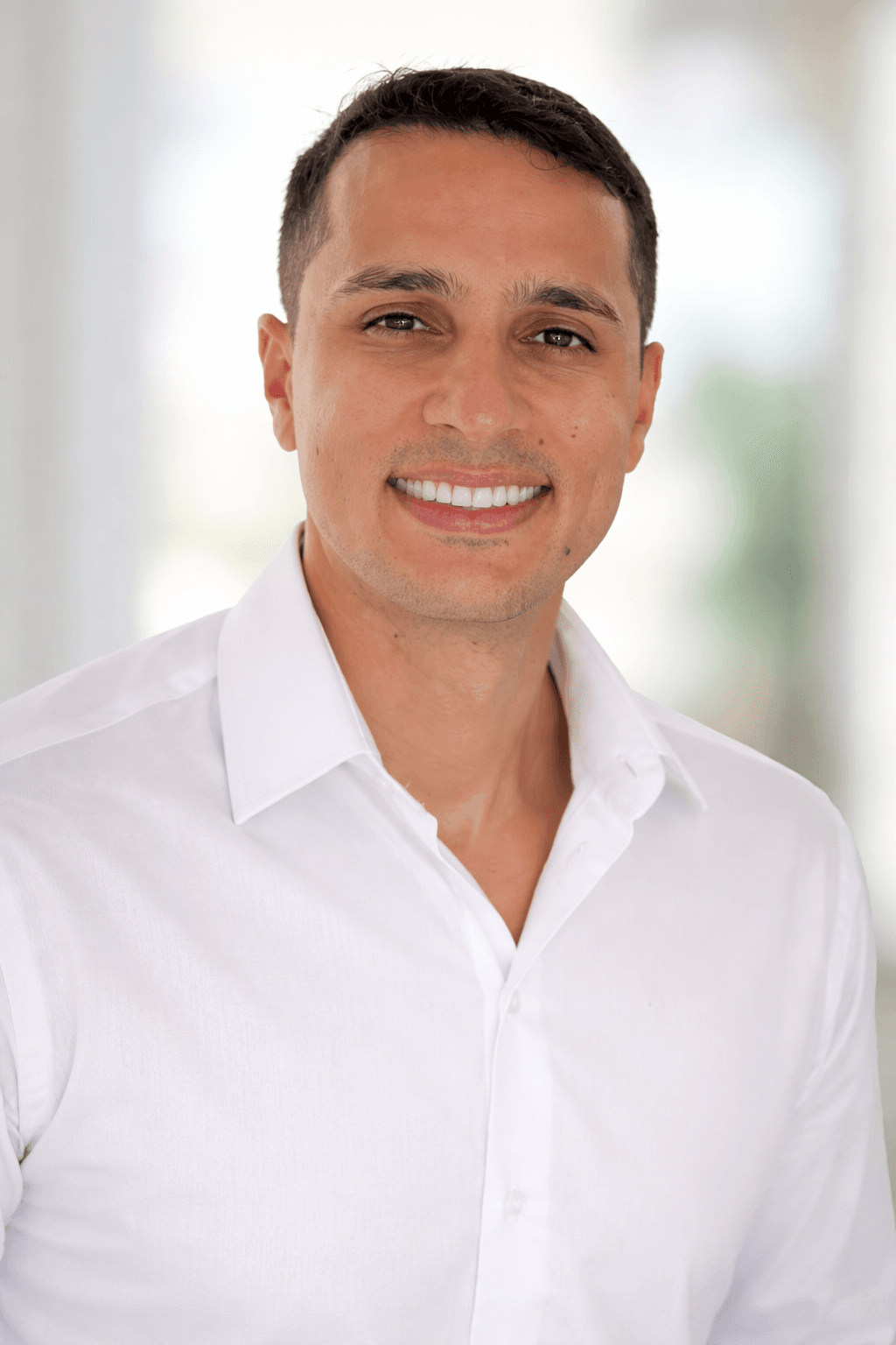 Smiling man with short dark hair wearing a white button-down shirt against a blurred background.