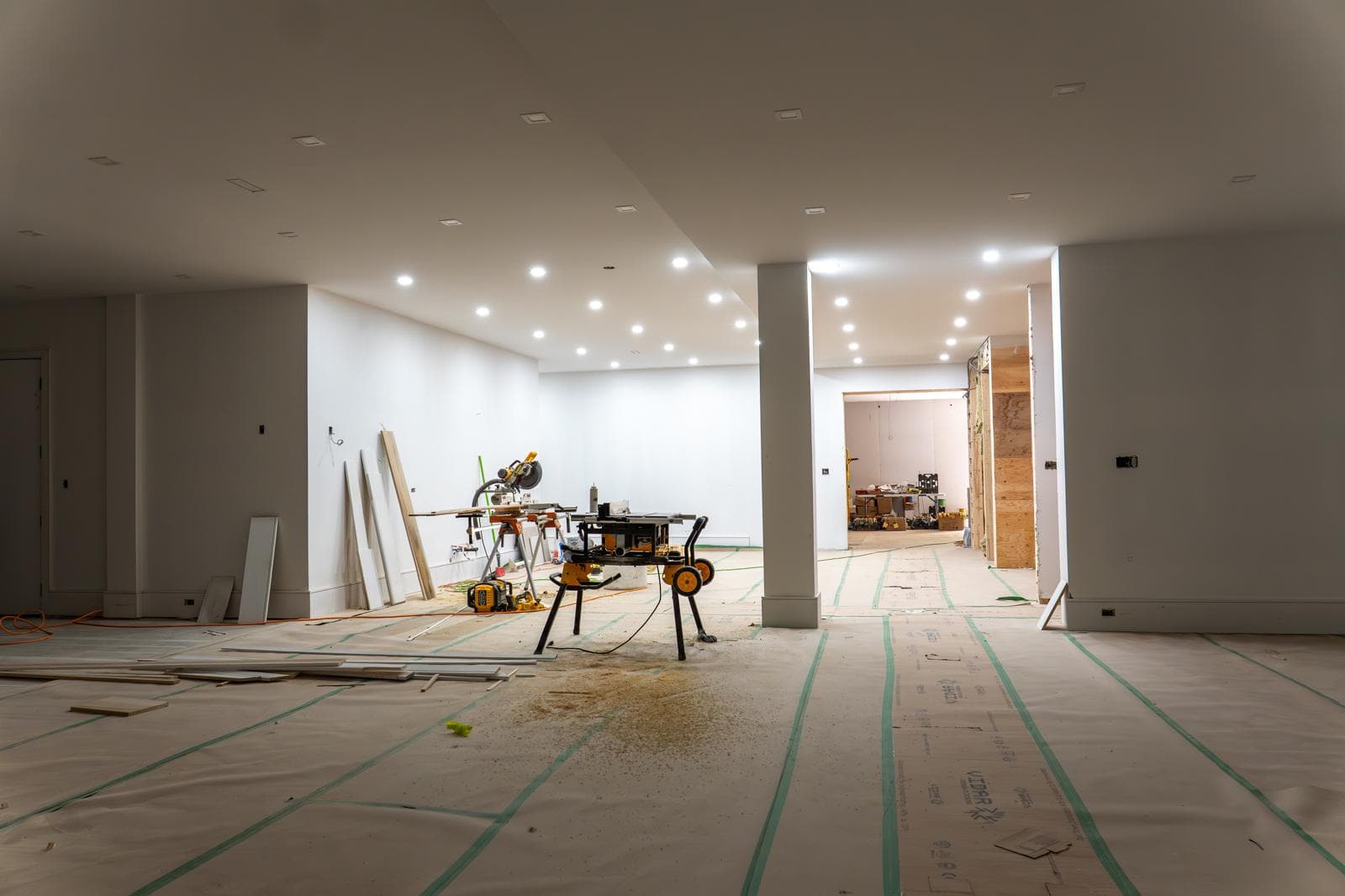 Brightly lit indoor renovation with white walls, recessed lighting, and power saws on covered floors.