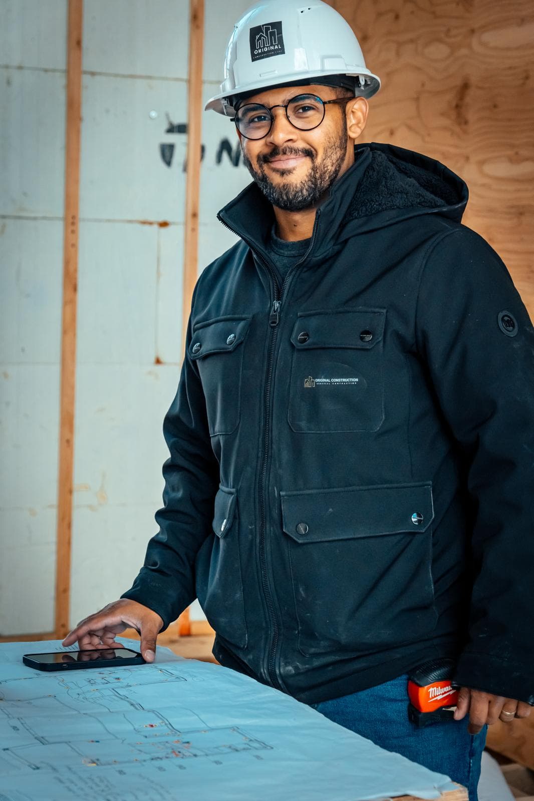Smiling man in a hard hat and jacket reviewing blueprints at a construction site.