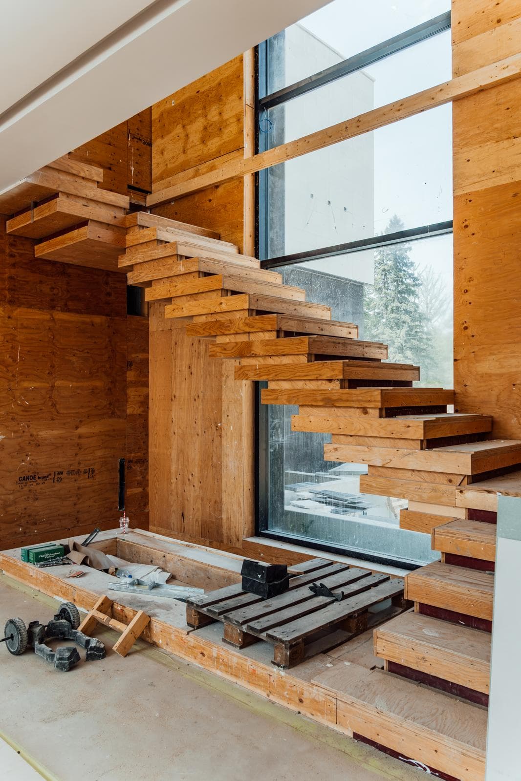 Modern floating wooden staircase under construction against plywood walls beside a large window.
