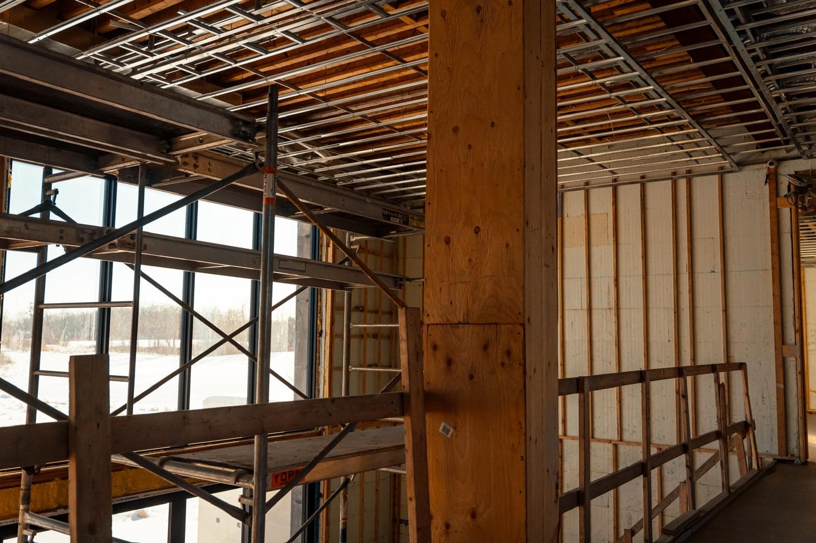 Interior construction site with scaffolding, wooden pillars, and exposed ceiling framing near large windows.