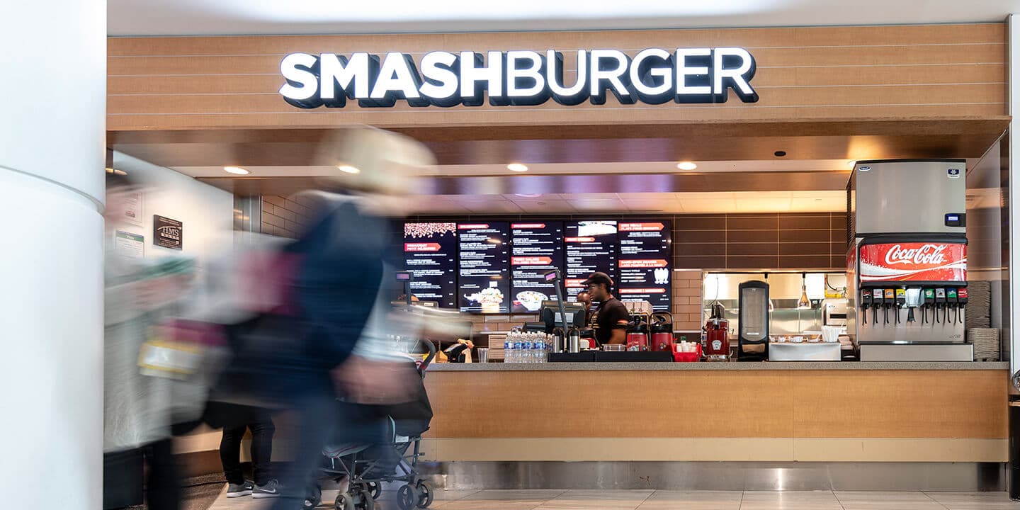 Smashburger counter with digital menus and a soda fountain, featuring blurred people in motion.